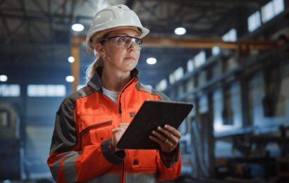 Professional Heavy Industry Engineer/Worker Wearing Safety Uniform and Hard Hat Uses Tablet Computer. Serious Successful Female Industrial Specialist Walking in a Metal Manufacture Warehouse.
