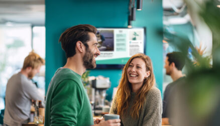 Colleagues laughing over coffee with a wallmounted digital screen in the background